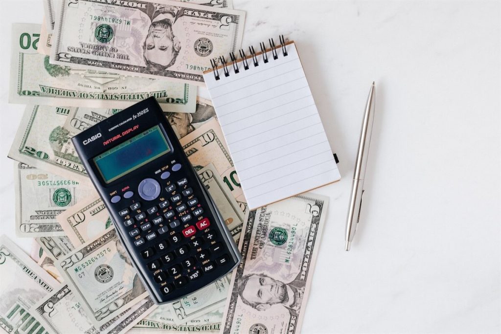 A calculator, pen, notepad and dollar bills on a table